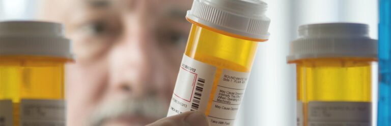 A person holding a prescription pill bottle, looking at the label. Several other pill bottles are in the foreground.