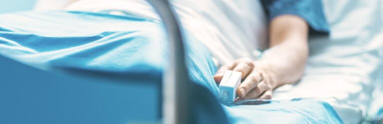 A person's hand resting on a hospital bed, wearing a pulse oximeter, signifying a medical setting where Medicare Part A covers care.