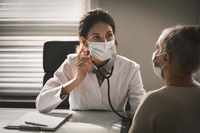 A healthcare professional wearing a mask is using a stethoscope to examine an elderly patient, who is also wearing a mask, in a well-lit examination room.