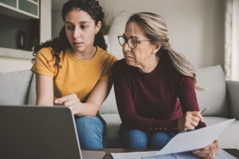 Older and younger woman looking at laptop and paperwork.
