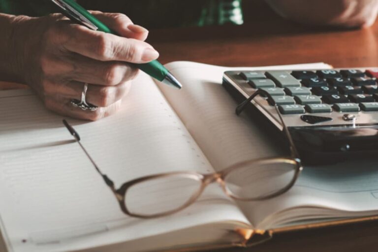 Glasses on a notebook, next to a calculator and a hand holding a pen.