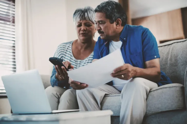 Couple on couch looking at bill and phone with laptop open, determining whether life insurance is worth it.